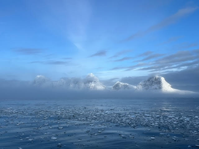 View from a ship of icy seas, with sunlit snow mountains in the distance.