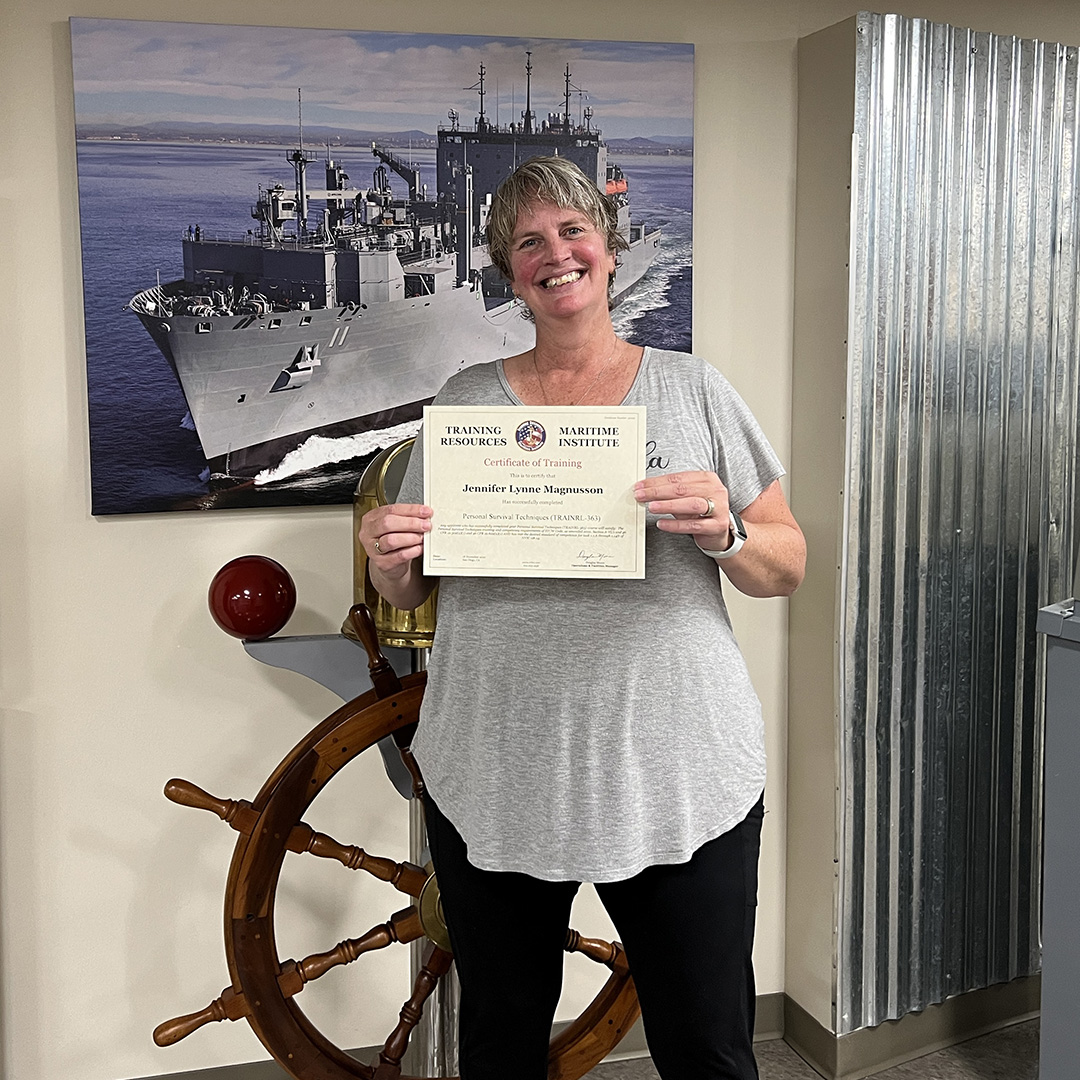 Woman stands in front of a ship's wheel holding a certificate