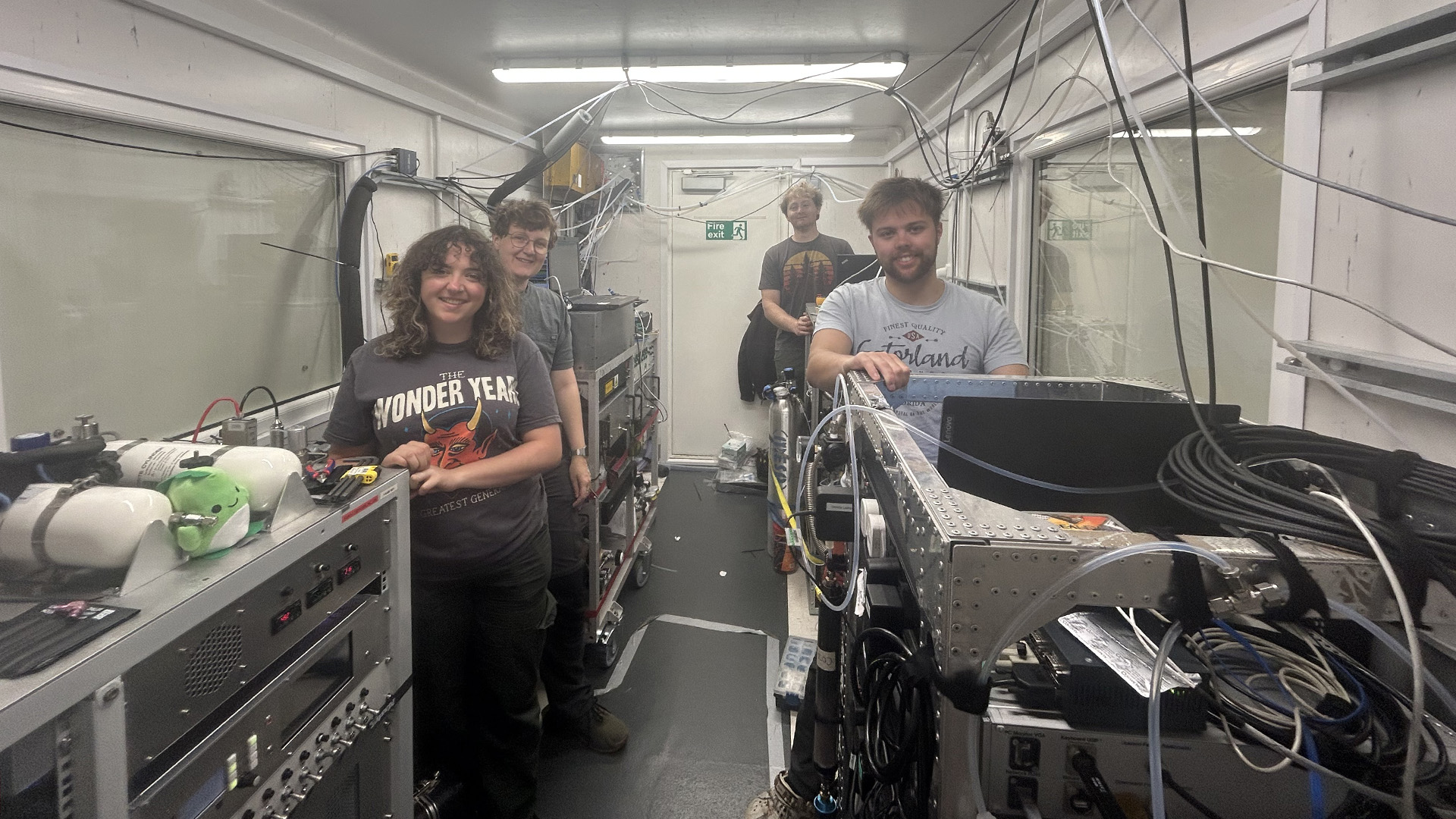 6.23 York Container 16x9 PhD students in the center container on the ship’s foredeck; from left to right: Tara Murphy with the gas-chromatograph mass-spectrometer to measure VOCs, Phin Petherick with two LIFs to measure NOx and SO2, Jared Novelly with a vintage mass spectrometer measuring SO2, and Jake Job with his instrument to measure ozone and NOx.
